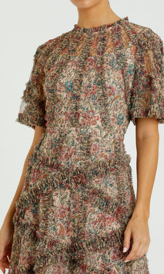 Someone wearing the Mac Duggal 8010 Short-Sleeve Wedding-Guest Dress, which features a tea-length cut, floral print, ruffled detailing, and sheer fabric on the upper chest and sleeves.