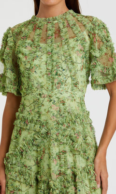 A woman wears the Mac Duggal 8010, a short-sleeve wedding-guest dress featuring green floral print, sheer fabric, and ruffled trim—perfect for parties or any special occasion.