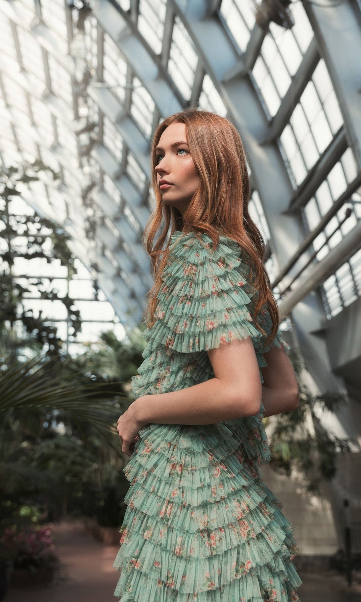 A woman with long auburn hair wears the Midi Wedding Guest Dress 8014 by Mac Duggal, a sage green floral dress with ruffled details, standing in a glass-walled greenhouse surrounded by lush plants. This semi-formal dress is perfect for any occasion.