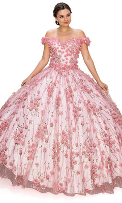 A woman gracefully poses in the Quinceanera Ball Gown 8021J—an off-the-shoulder pink dress featuring 3D floral details and a lace-up corset bodice, elegantly holding the sides of her gown.