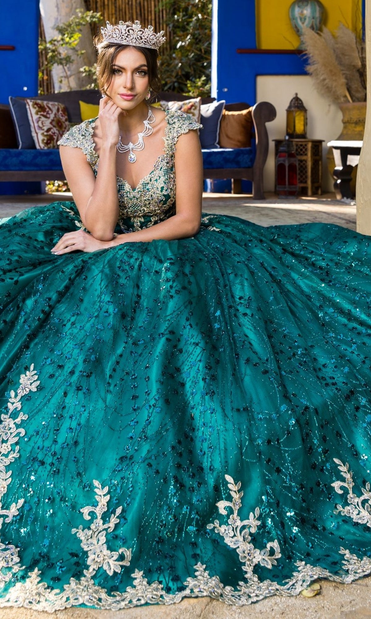 Wearing the Quinceanera Ball Gown 8024J with gold lace embroidery, a woman in a jeweled crown sits indoors on the floor, surrounded by elegant decor and a couch in the background.
