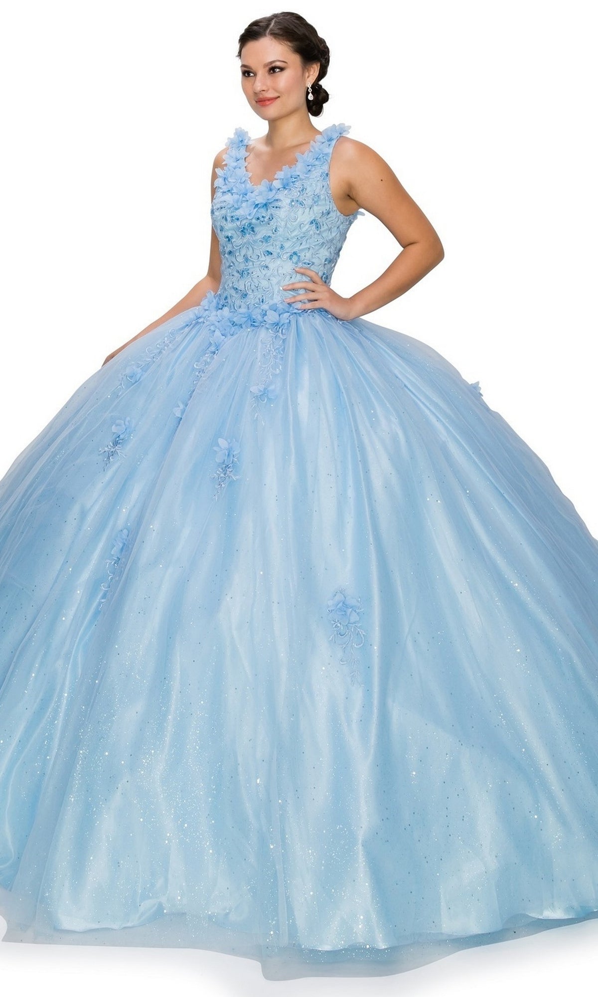 A woman wears the Quinceanera Ball Gown 8025J, featuring a light blue color, floral appliques, and a full glitter tulle skirt, standing against a plain white background.