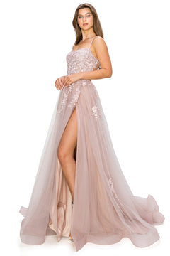A woman stands against a white background wearing the Floral-Embroidered Long Tulle Prom Dress 8031J, a blush pink, floor-length gown with floral lace details, spaghetti straps, and a high leg slit.