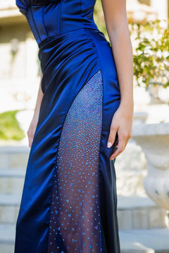 Someone stands outdoors near stone steps and a planter, wearing the Sheer Side-Slit Long Corset Prom Dress 8037J—a blue satin gown with a sheer, rhinestone-embellished panel.