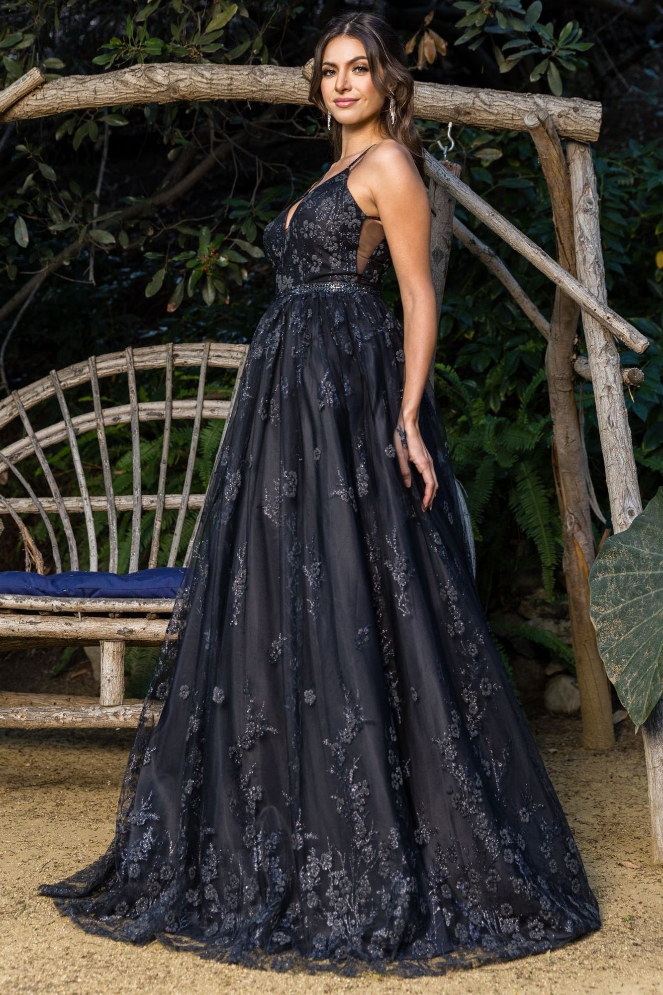 A woman stands outdoors in the Corset-Back Long Glitter Prom Ball Gown 8039J—an elegant, sleeveless, floor-length black dress with floral patterns—next to a rustic wooden bench and lush greenery.