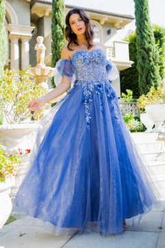 A woman in a blue Puff-Sleeve Long Sweetheart Prom Ball Gown 8042J stands outdoors near a stone fountain and lush greenery.