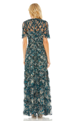 A woman stands facing away, wearing the Mac Duggal 8043 long formal dress, featuring short sleeves and a detailed blue, green, and beige floral pattern.