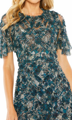 A woman models the Mac Duggal 8043 Long Formal Dress—a teal, textured short-sleeve dress with floral pattern and sheer panels—shown from shoulders to mid-thigh.