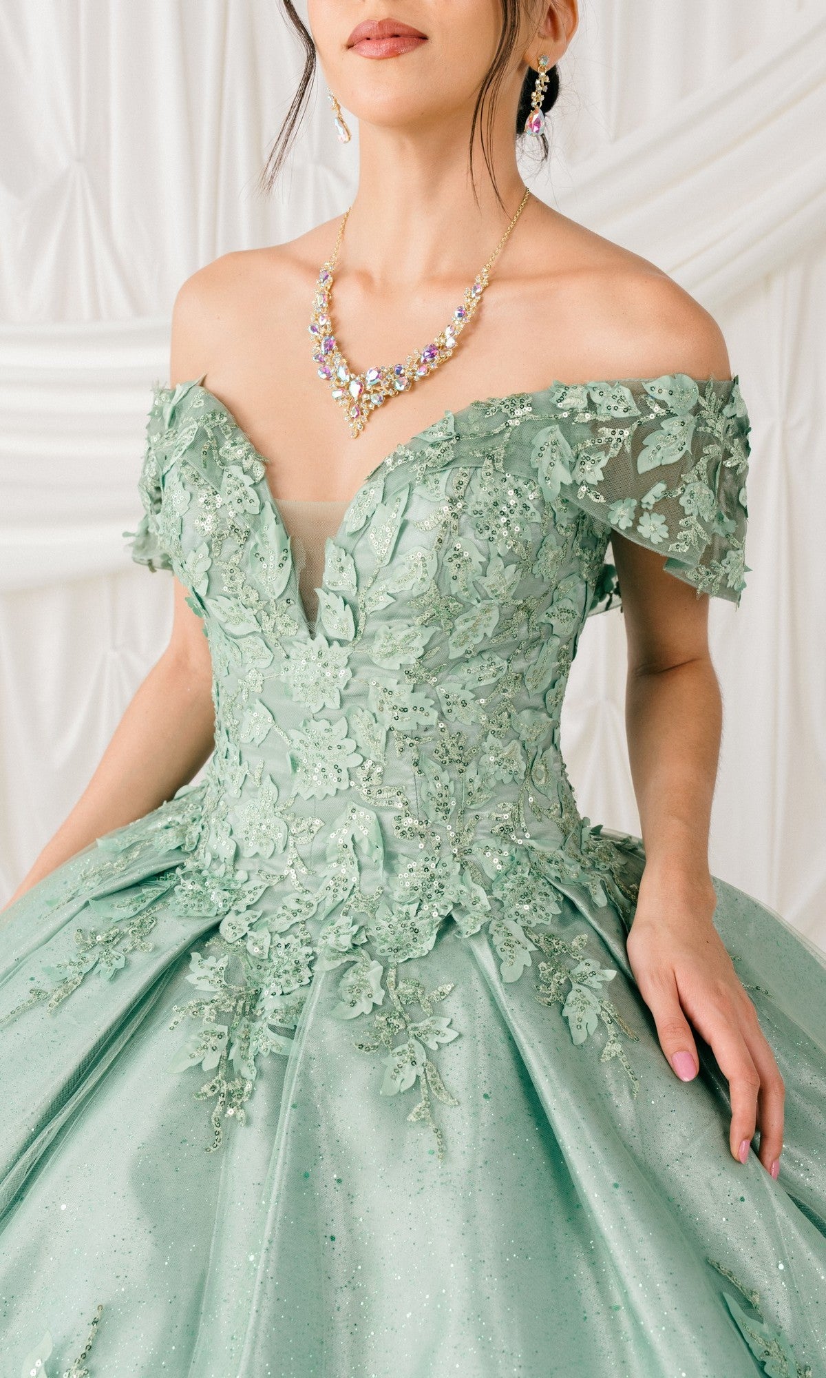A woman wearing the Quinceanera Ball Gown 8045J with glitter lace embroidery and 3D floral detail, plus a beaded necklace and earrings, stands before a white draped backdrop.
