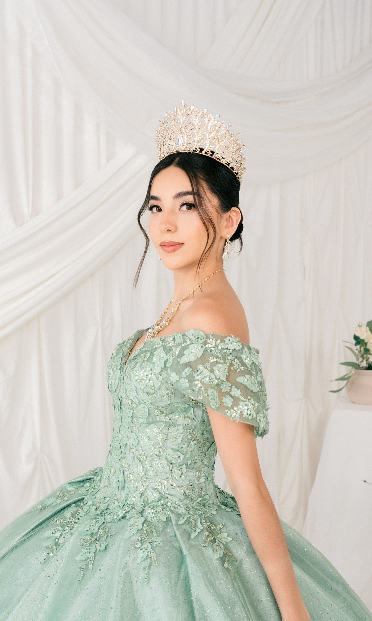 A young woman wears the Quinceanera Ball Gown 8045J in light green, featuring off-the-shoulder glitter lace embroidery and a jeweled crown, posing before white draped fabric.