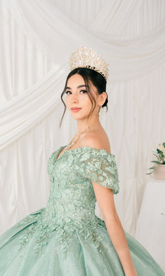 A young woman wears the Quinceanera Ball Gown 8045J in light green, featuring off-the-shoulder glitter lace embroidery and a jeweled crown, posing before white draped fabric.