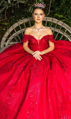 A woman in the Quinceanera Ball Gown 8045J, featuring red off-the-shoulder glitter lace embroidery and a jeweled crown, sits on a round wooden bench with greenery behind her.
