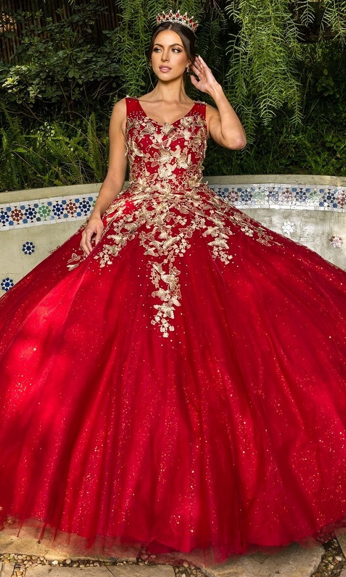A woman wearing the Quinceanera Ball Gown 8046J, featuring metallic embroidery and a tiara, stands in front of a tiled fountain surrounded by lush greenery.