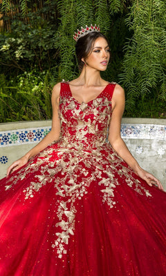 A woman wearing the Quinceanera Ball Gown 8046J, featuring glitter tulle and gold metallic embroidery, with a red jeweled tiara, seated outdoors in front of lush greenery.