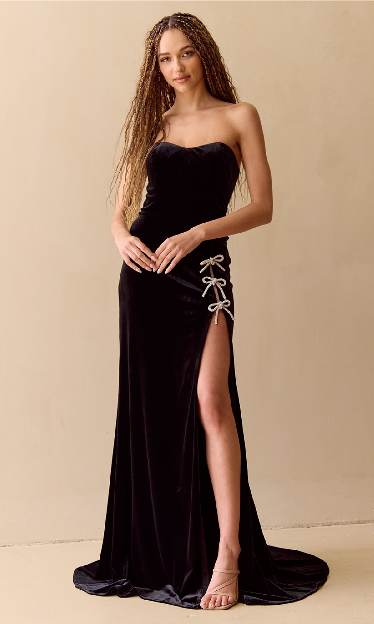 A woman wearing the Amelia Couture 8047, a long strapless velvet prom dress with a thigh-high slit and bow details, stands against a plain beige background.