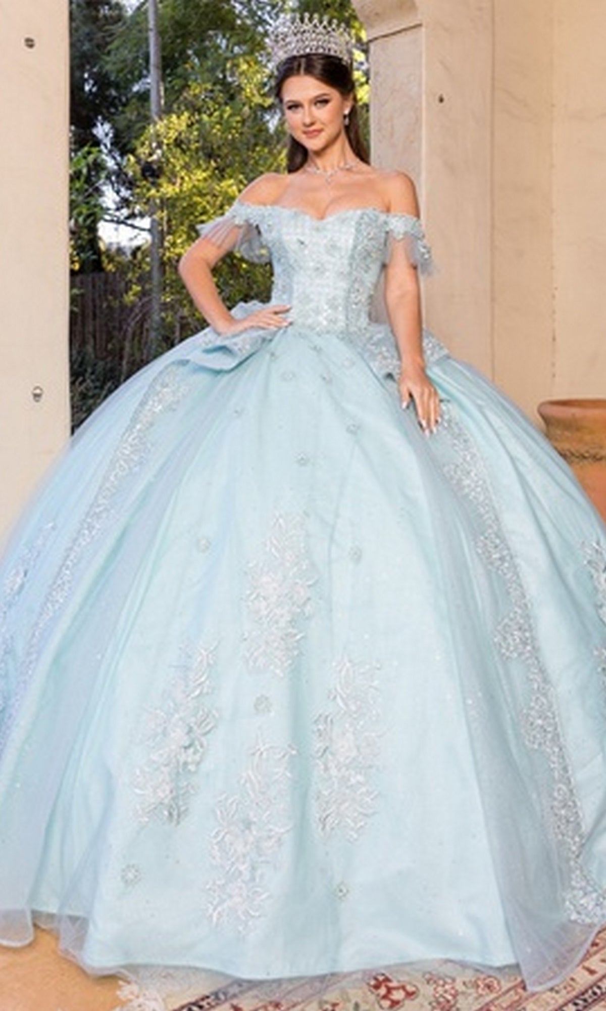 A woman stands on a porch wearing the Quinceanera Dress 8055J by Cinderella Couture, a light blue off-the-shoulder ball gown with lace embroidery and a tiara.