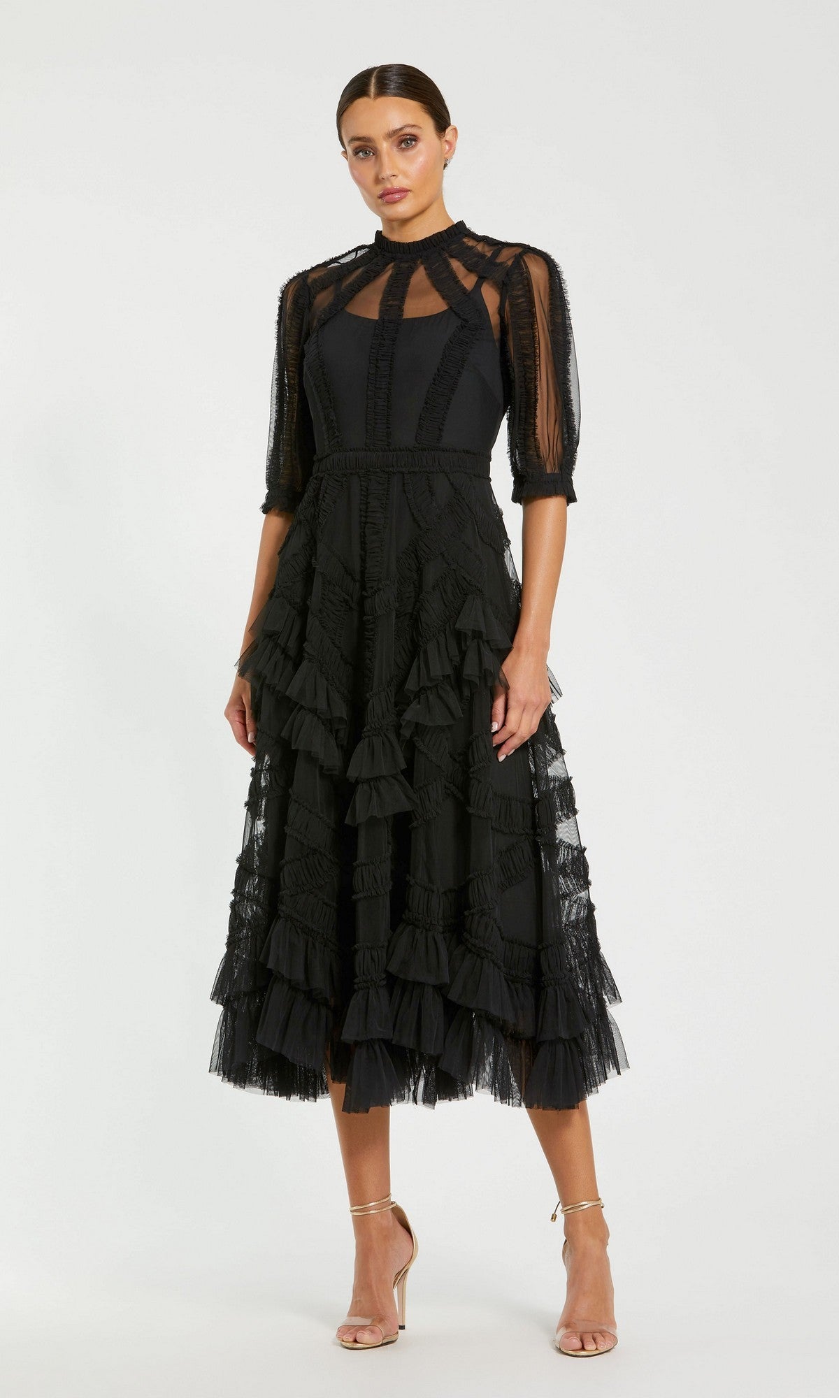 A woman stands against a plain background wearing the black Mac Duggal 8058 Ruffled Midi Wedding-Guest Dress, featuring sheer sleeves, a high neck, layered ruffles, and strappy nude heels.