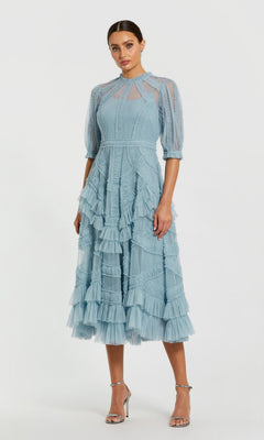 A woman models the Mac Duggal 8058 Ruffled Midi Wedding-Guest Dress in French Blue, featuring sheer ruffled sleeves, ruffle details, and a high neckline, standing against a plain white background.