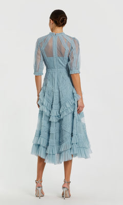 A woman stands with her back to the camera in a Mac Duggal 8058 Ruffled Midi Wedding-Guest Dress, light blue with sheer panels and ruffled layers, paired with heeled sandals; her hair is styled in a neat bun.