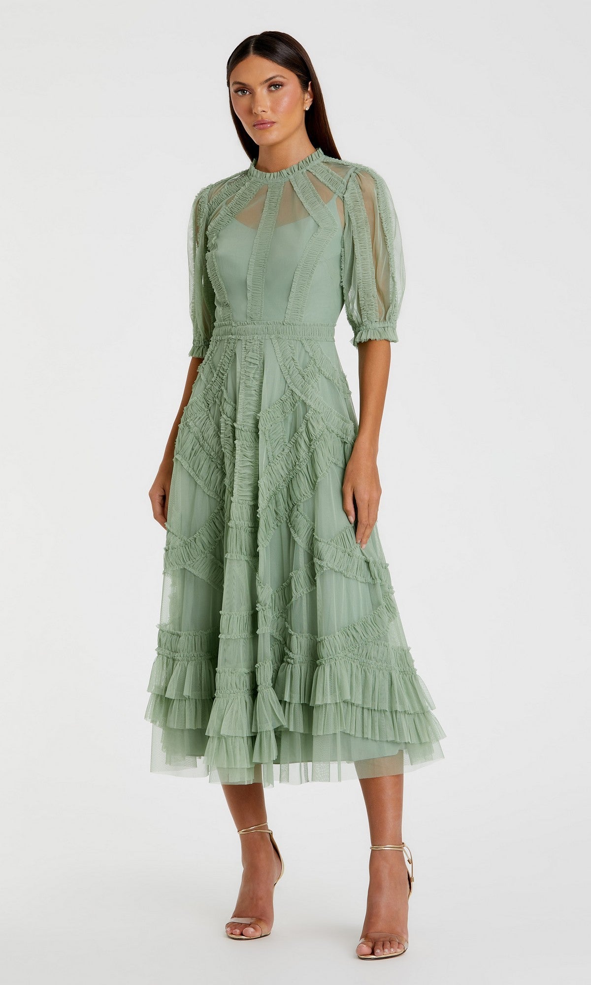 A woman stands against a plain background wearing the Mac Duggal 8058 Ruffled Midi Wedding-Guest Dress in jade, featuring sheer sleeves and ruffled skirt layers, styled with strappy gold heels.