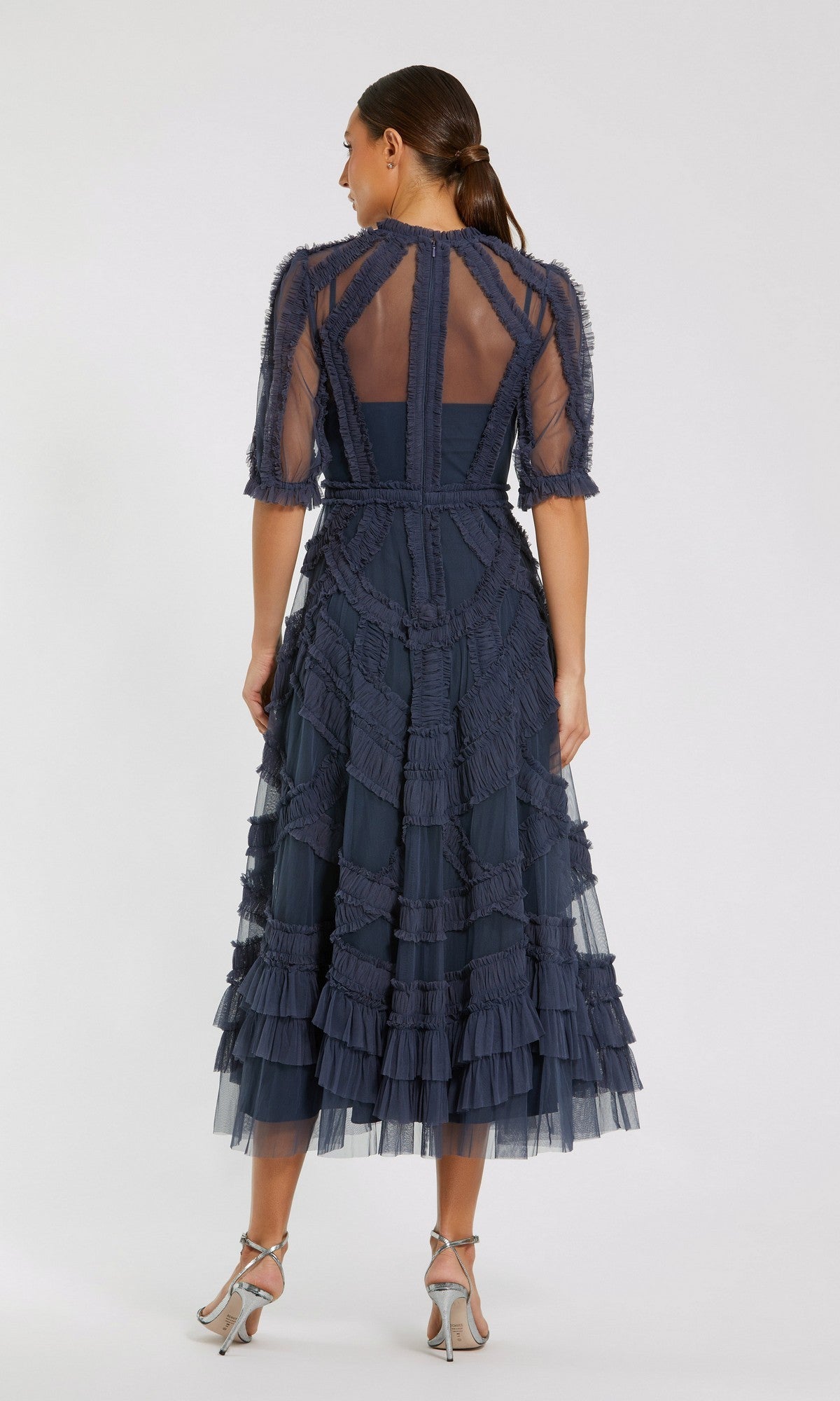 A woman faces away from the camera, wearing the Mac Duggal 8058 Ruffled Midi Wedding-Guest Dress in navy blue with ruffled sleeves and sheer accents, styled with silver high-heeled sandals.