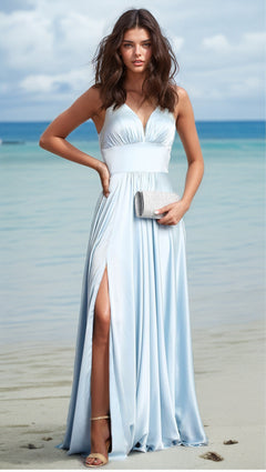 A woman stands on the beach holding a silver clutch, wearing the Empire-Waist Long A-Line Prom Dress: Chicas C242—a light blue, sleeveless, floor-length gown with a thigh-high slit—with the ocean in the background.