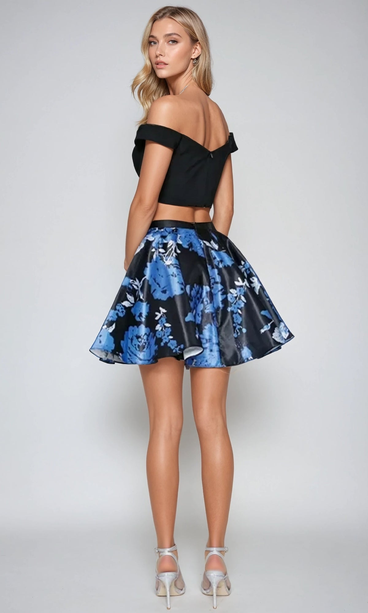 A woman stands facing away, wearing the Juliet 805 short two-piece hoco dress—an off-the-shoulder black crop top paired with a flared blue floral A-line skirt and high heels, against a plain light background.