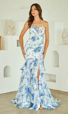 A woman stands indoors by a white wall, modeling the Strapless Print Prom Gown: Amelia Couture 8060—featuring blue and white florals, ruffle accents, and a thigh-high slit.
