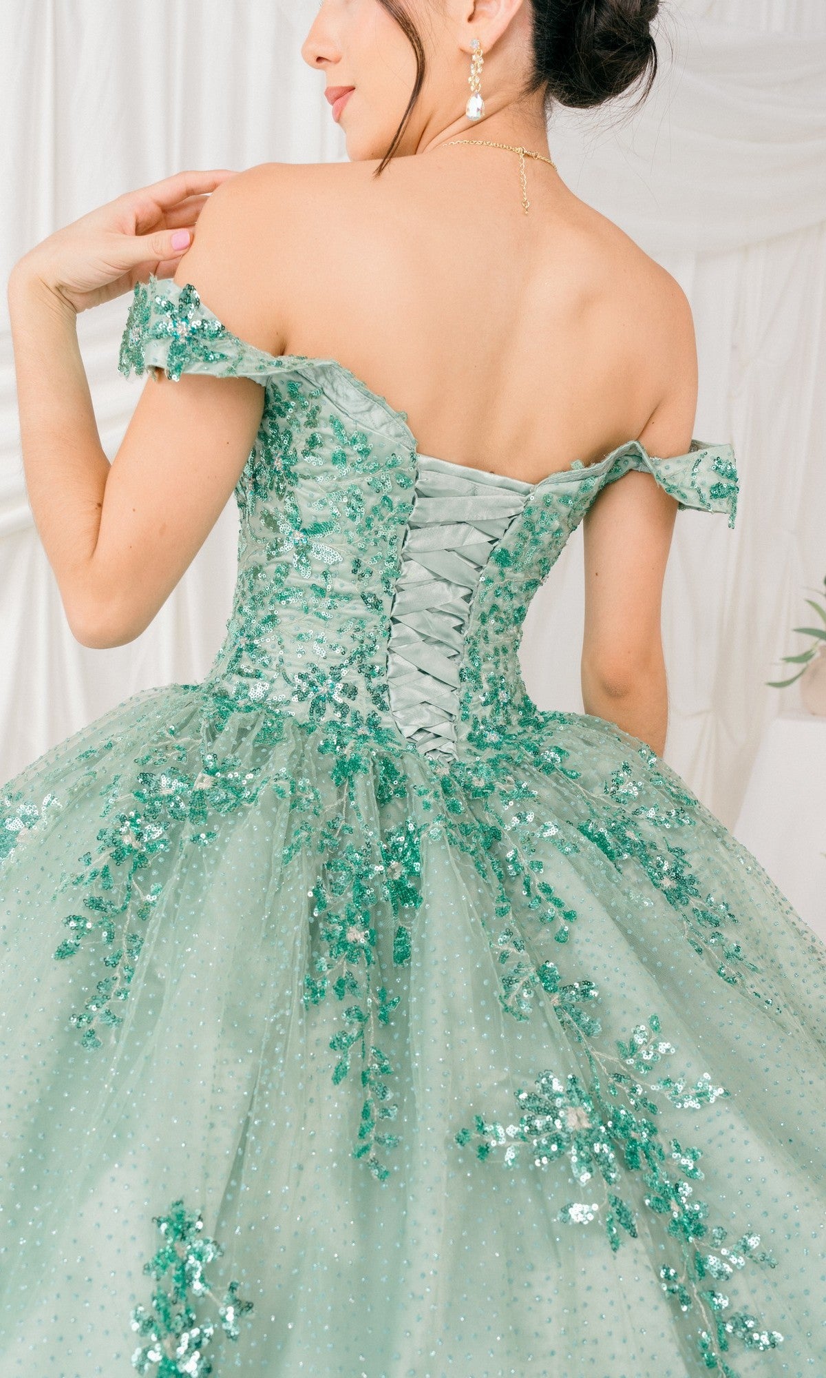 Model shown from behind wearing the Quinceanera Ball Gown 8060J in light green, featuring off-the-shoulder design, sequin and floral embellishments, and a lace-up back, set against a white fabric backdrop.