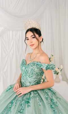 A young woman wearing the Quinceanera Ball Gown 8060J— a green sequin off-the-shoulder dress with a lace-up back and crown— poses indoors before white drapery and floral arrangements.