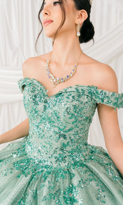 A woman wears the Quinceanera Ball Gown 8060J—an off-shoulder, green ball gown with embossed tulle and lace-up back—paired with jeweled necklace and earrings, standing before a white, draped backdrop.