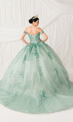 A woman wearing a tiara poses with her back to the camera in the Quinceanera Ball Gown 8060J, an off-the-shoulder pastel green embossed tulle dress with floral embellishments and a lace-up back, set against a white draped backdrop.