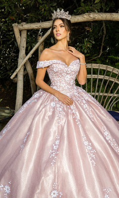 A woman wearing a silver tiara and the Quinceanera Ball Gown 8060J—an off-the-shoulder dress with pink embossed tulle and floral embroidery—stands before wooden branches and foliage.