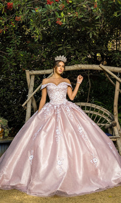 A woman in the Quinceanera Ball Gown 8060J, a sparkly off-the-shoulder dress with embossed tulle and a jeweled crown, stands before a wooden arch decorated with greenery and red flowers.