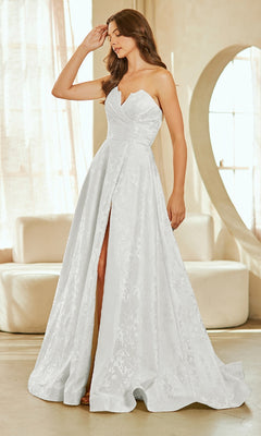 A woman wears the Amelia Couture 8061 Textured Strapless Prom Gown with a sweetheart neckline and front slit, standing indoors near a round mirror and sofa.
