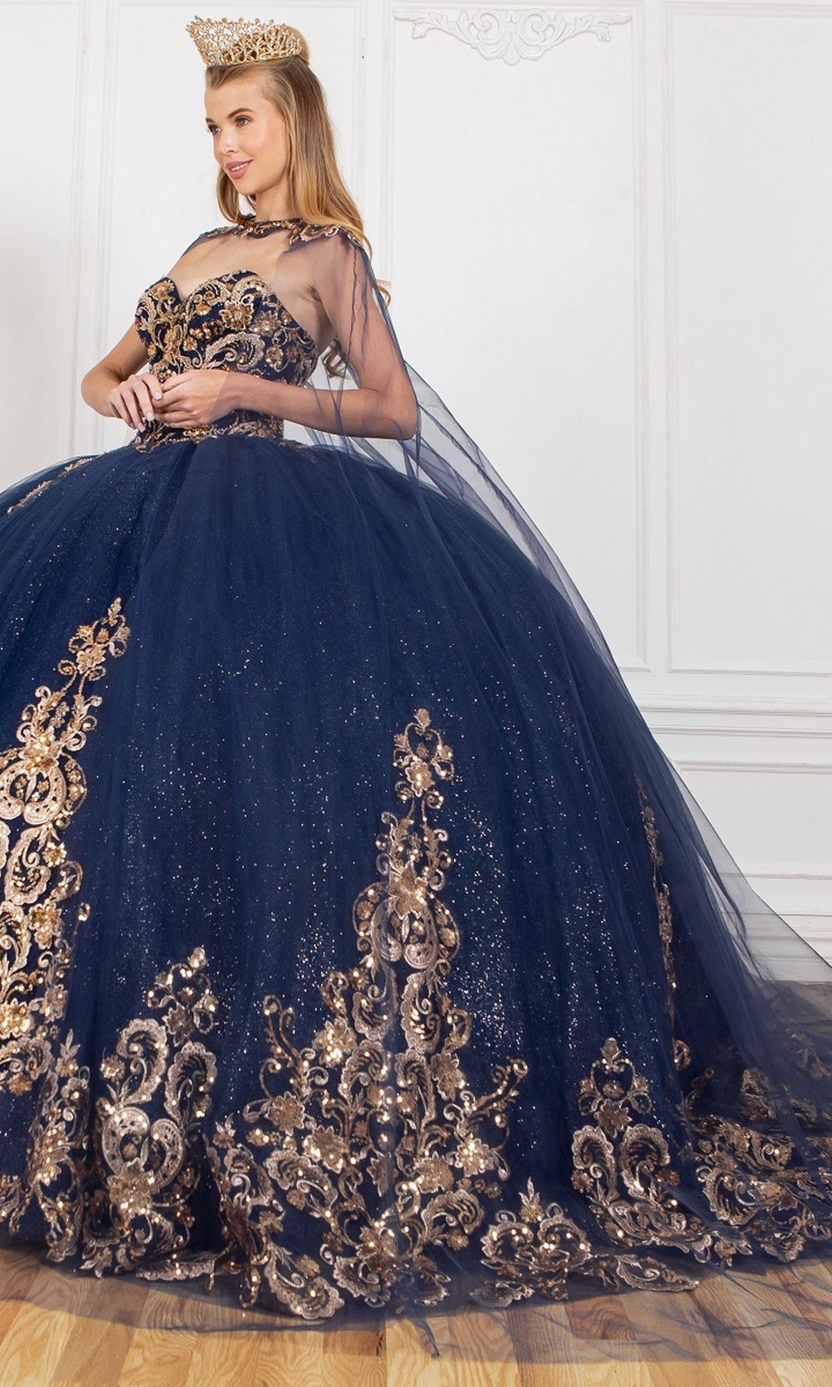 A woman in the Quinceanera Ball Gown 8063J—a dark blue strapless sweetheart dress with gold embroidery and glitter—wears a gold crown and stands against a white wall.