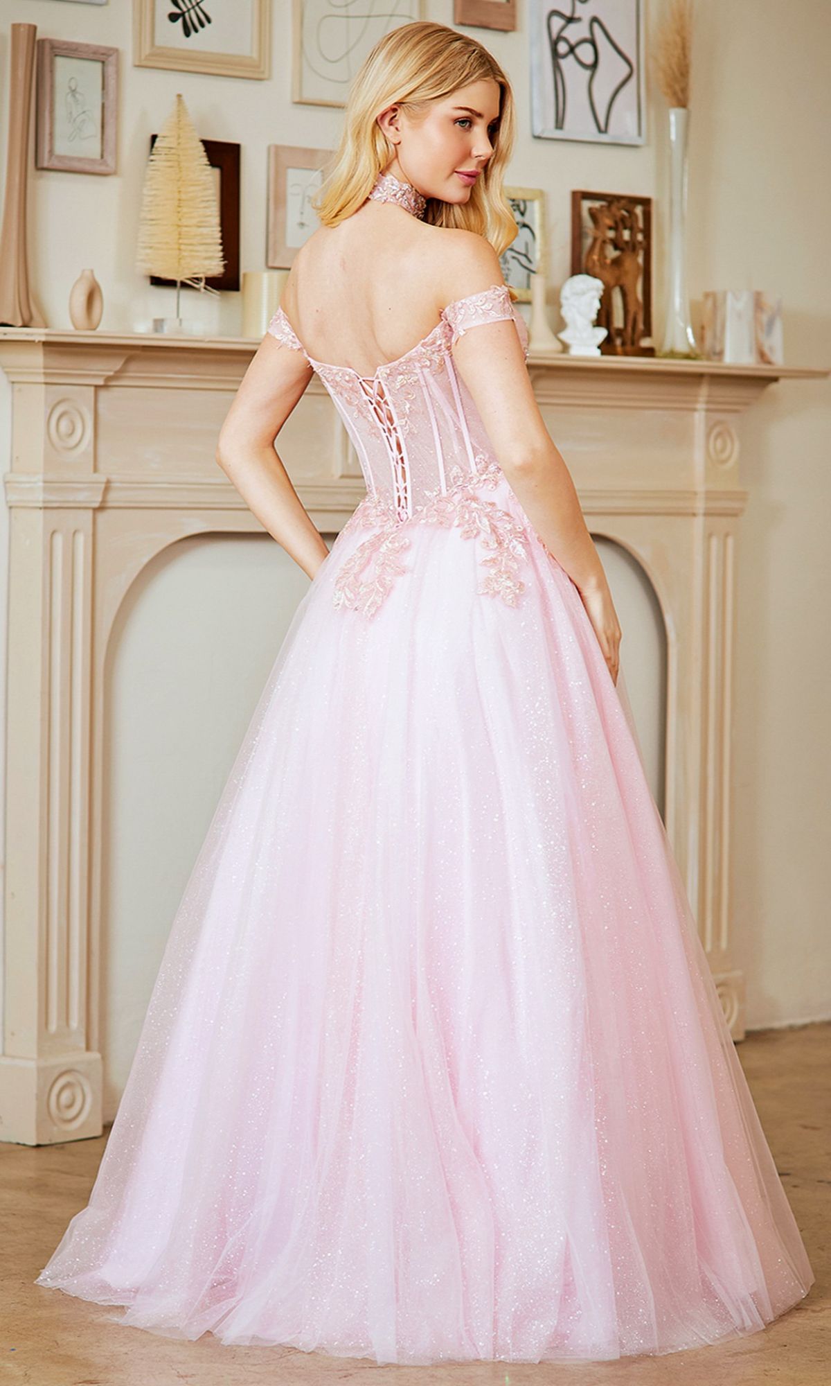 A woman in the Amelia Couture 8064 long prom dress, featuring light pink off-the-shoulder styling and a lace-up back, stands before a decorated mantelpiece, facing away from the camera.
