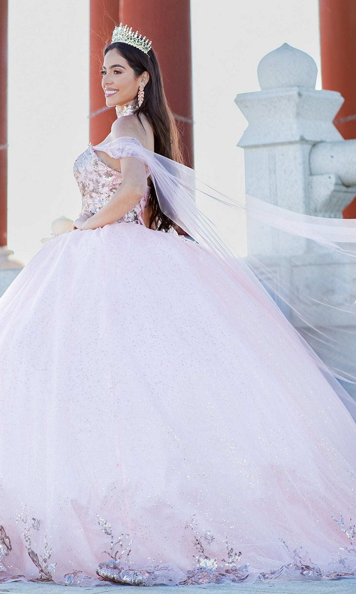A young woman poses outdoors beside red columns and stone structures, wearing the Quinceanera Ball Gown 8064J—an off-the-shoulder dress with a sparkling light pink skirt and a crown.