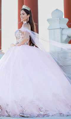 A young woman poses outdoors beside red columns and stone structures, wearing the Quinceanera Ball Gown 8064J—an off-the-shoulder dress with a sparkling light pink skirt and a crown.
