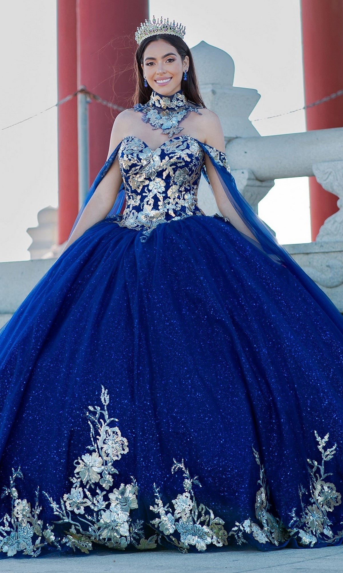 A young woman stands outdoors, smiling at the camera in the Quinceanera Ball Gown 8064J with an off-the-shoulder design, jeweled crown, and silver floral embellishments.