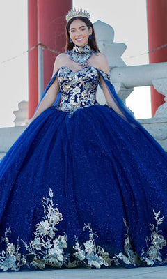 A young woman stands outdoors, smiling at the camera in the Quinceanera Ball Gown 8064J with an off-the-shoulder design, jeweled crown, and silver floral embellishments.