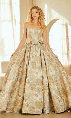 A woman stands indoors wearing the Amelia Couture 8068 Lace-Up Print Prom Ball Gown—a strapless, long floral dress in pale blue and gold—gracefully holding the sides of her gown.