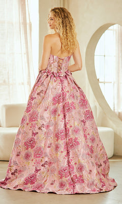 A woman with curly blonde hair stands in a sunlit room wearing the Lace-Up Print Prom Ball Gown: Amelia Couture 8068, a strapless pink floral ball gown featuring a lace-up back and bow.