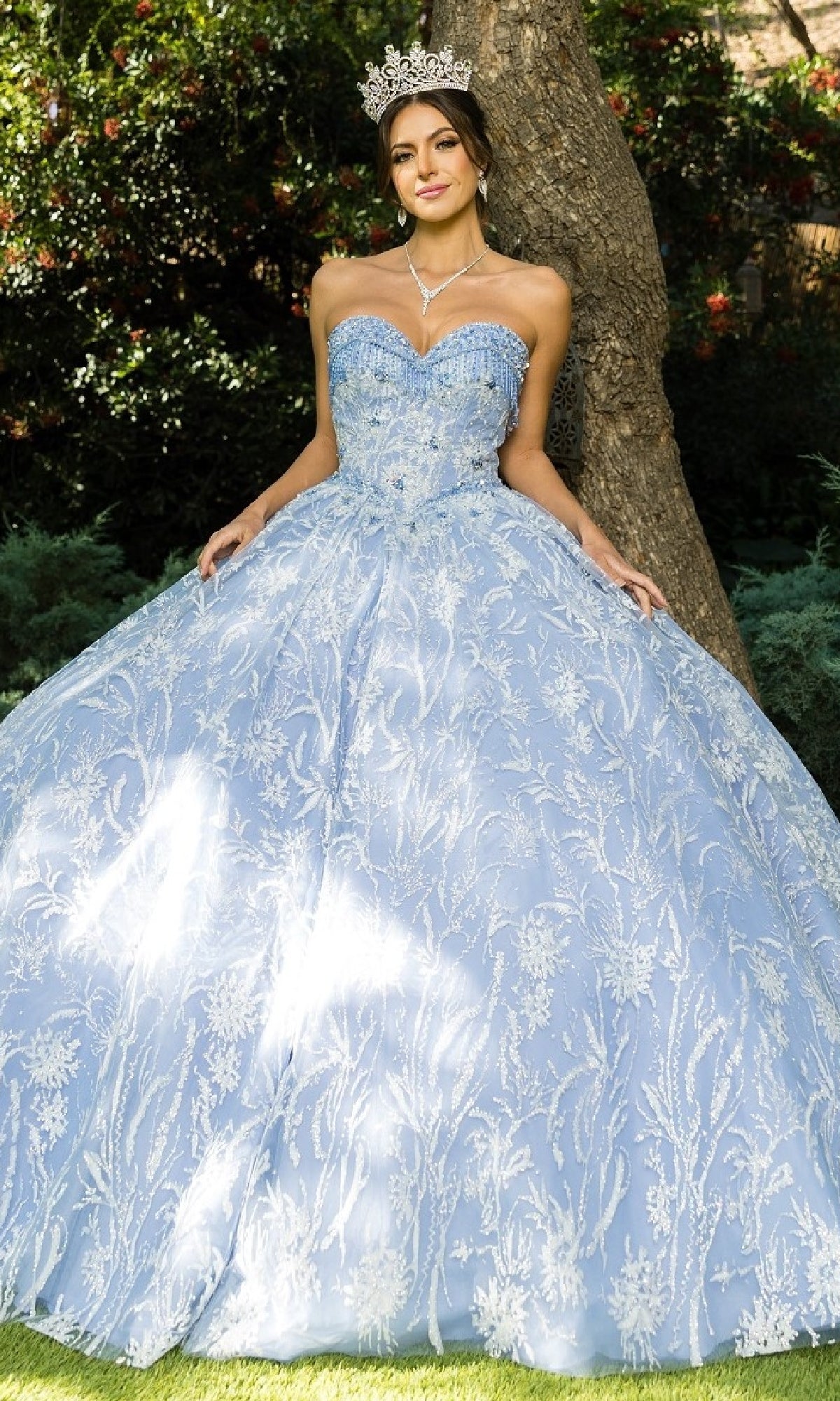 A woman stands outdoors in front of a tree wearing the Cinderella Couture 8071J quinceanera dress, a sparkling, strapless ball gown with beaded fringe and a tiara.