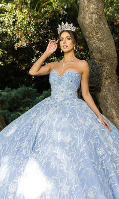 A woman poses outdoors near a tree in the Cinderella Couture 8071J quinceanera dress, a light blue strapless ball gown with beaded fringe, and wears a jeweled crown.