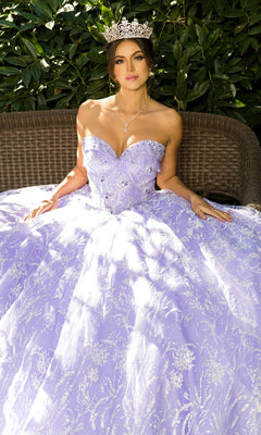 A woman in the Cinderella Couture 8071J quinceanera dress, featuring intricate embroidery and beaded fringe, wears a silver tiara as she sits on a wicker bench before lush green foliage.