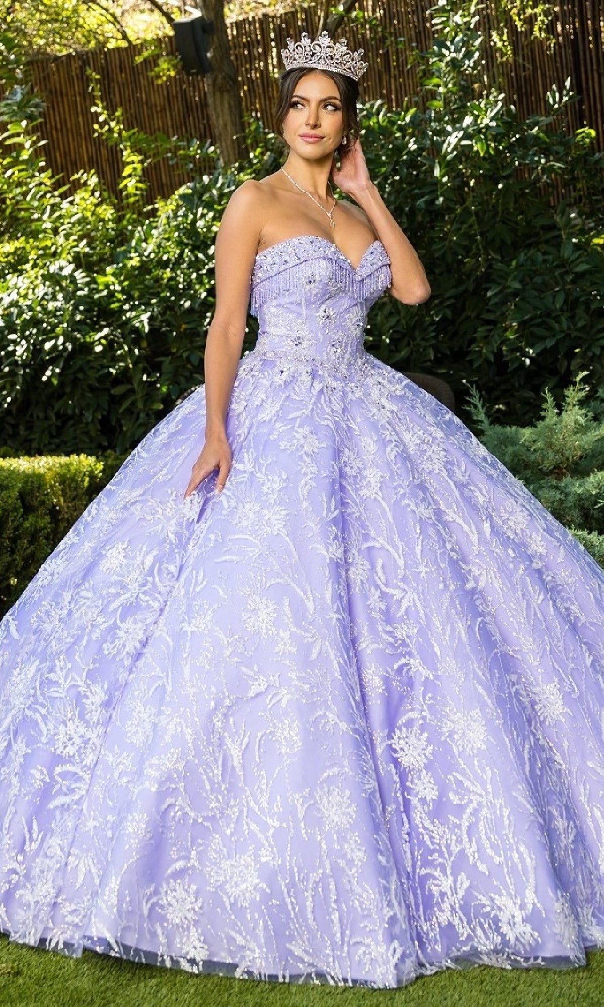 A woman wearing the Cinderella Couture 8071J sparkling lavender strapless quinceanera dress with beaded fringe and a tiara stands outdoors in a garden.