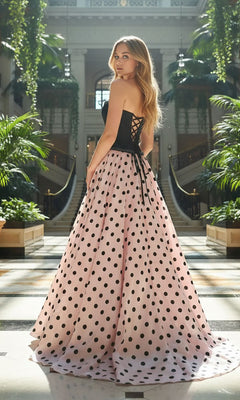 A woman in the Amelia Couture 8071 long prom dress stands in an elegant indoor garden, looking over her shoulder. The strapless black and pink polka dot a-line gown creates a striking, graceful silhouette.