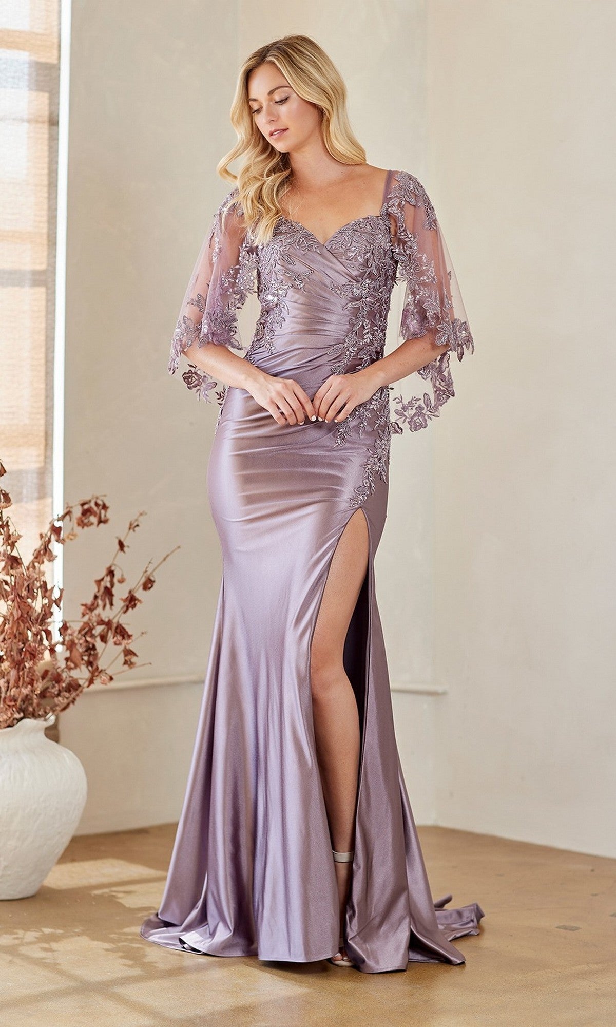 A woman in the Amelia Couture 8073 lilac satin long formal dress with embellished lace, sheer sleeves, and a high leg slit stands indoors near a white vase holding dried branches.