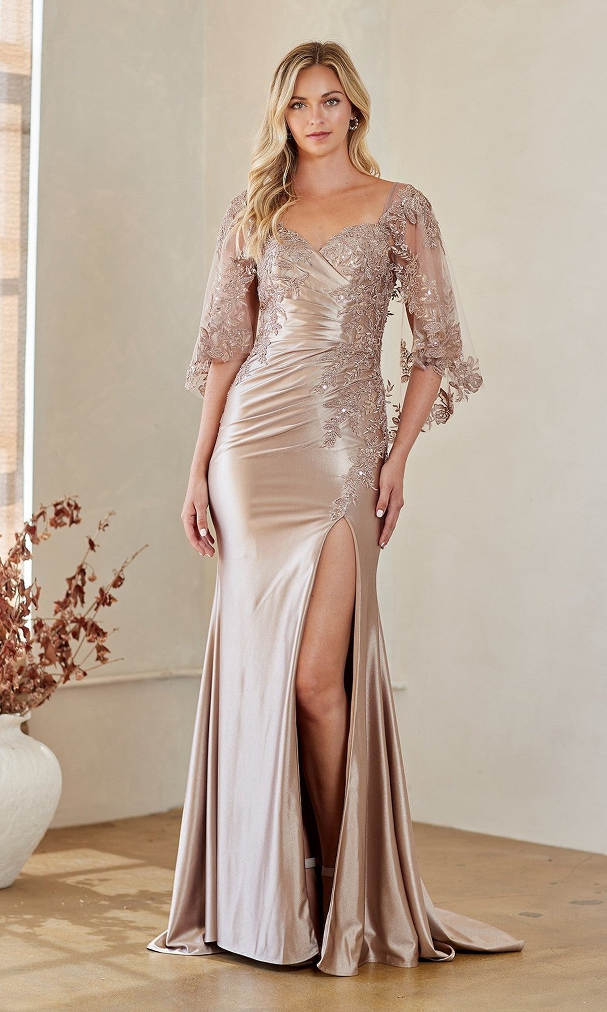 A woman in the Amelia Couture 8073 champagne satin gown with lace embroidery, elbow-length sleeves, and a high thigh slit stands indoors beside a white vase filled with dried branches.